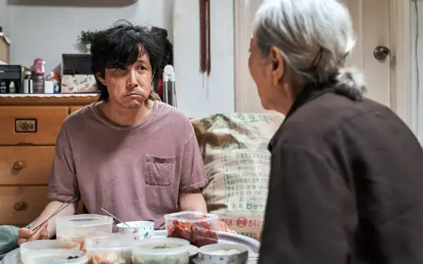 A scene from the TV show Squid Game featuring Lee Jung-jae, sitting at a table with dishes, engaged in a conversation with an elderly woman. Vibrant 4K Ultra HD wallpaper.