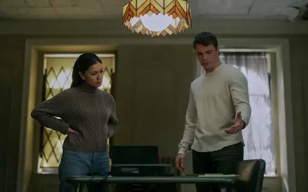 Gabriel Basso and Luciane Buchanan from the TV show The Night Agent engage in a tense conversation in this HD wallpaper featuring a dimly lit room and vintage lamp.