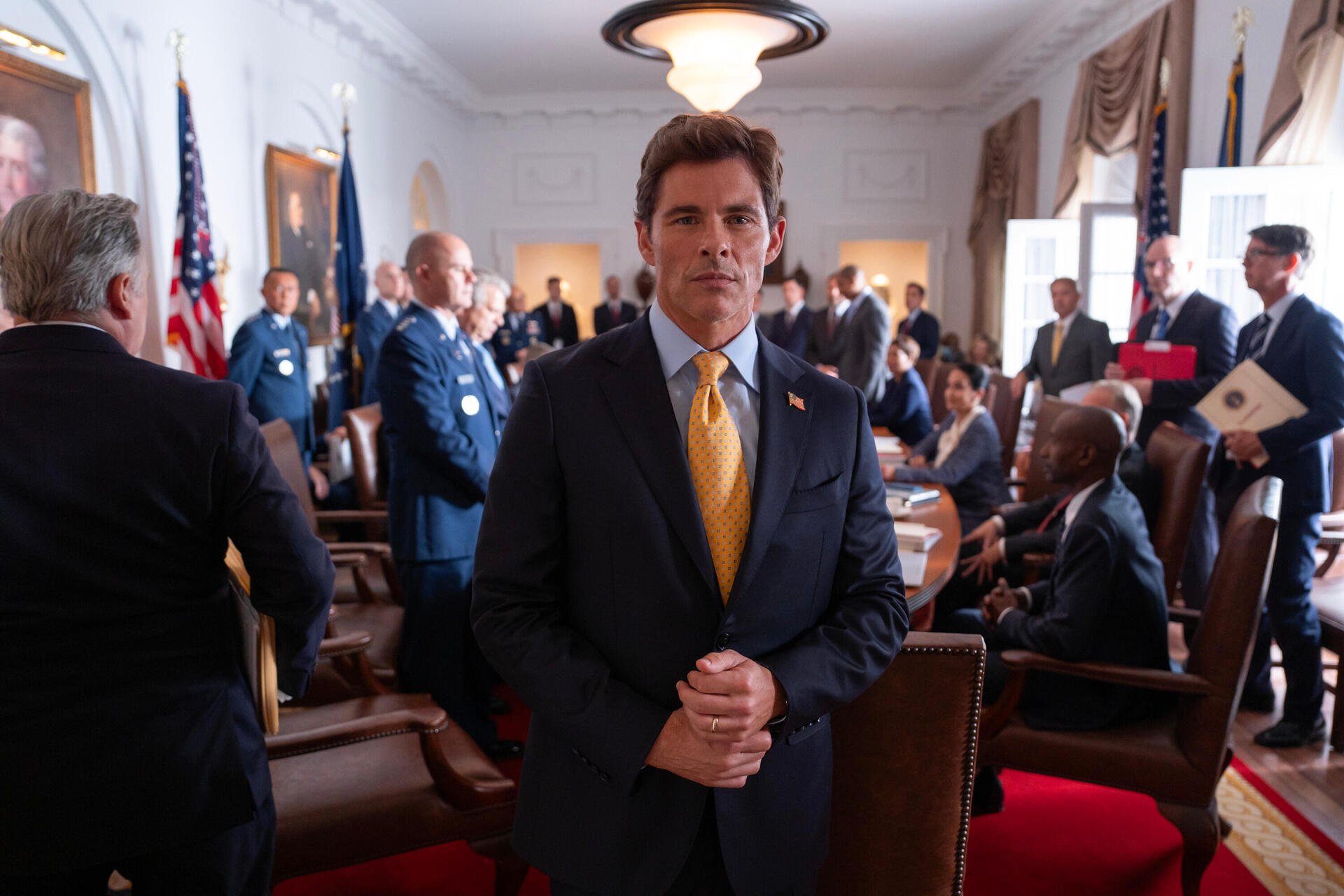 James Marsden stands in a formal setting from the TV show Paradise (2025), surrounded by a group of people engaged in discussion in an elegant room.