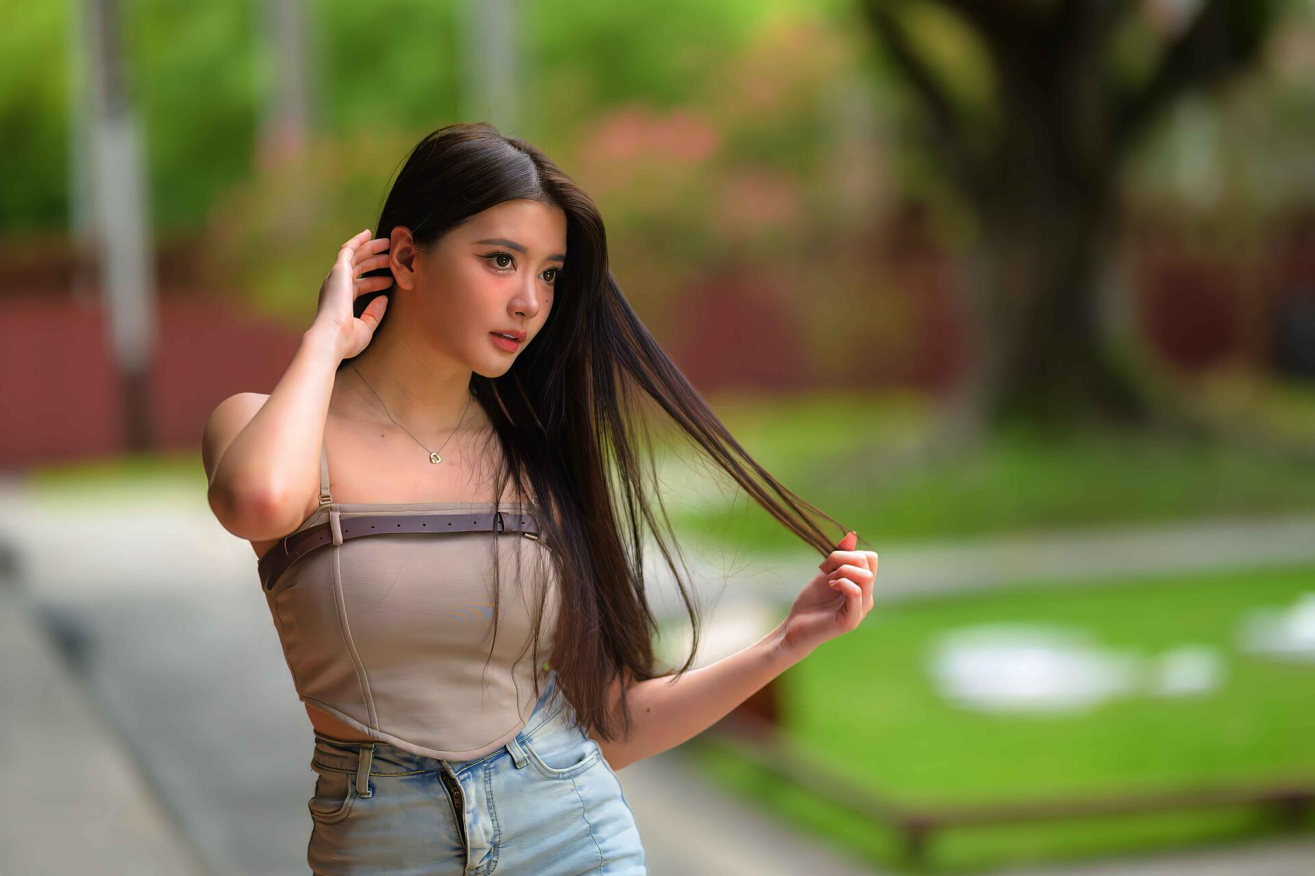 8K Ultra HD PC wallpaper: young woman in a tube top and jeans brushes her long hair, standing against a softly blurred green park background.
