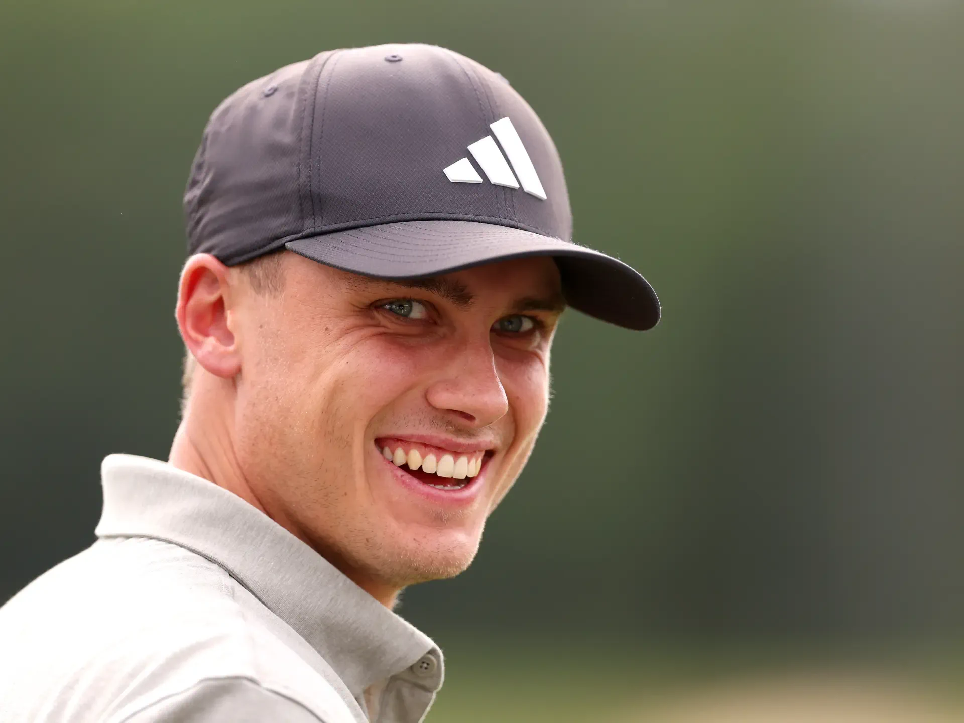 A close-up of Ludvig Åberg smiling while wearing an Adidas cap, set against a blurred green background. This HD image captures the energy and spirit of sports and golf.