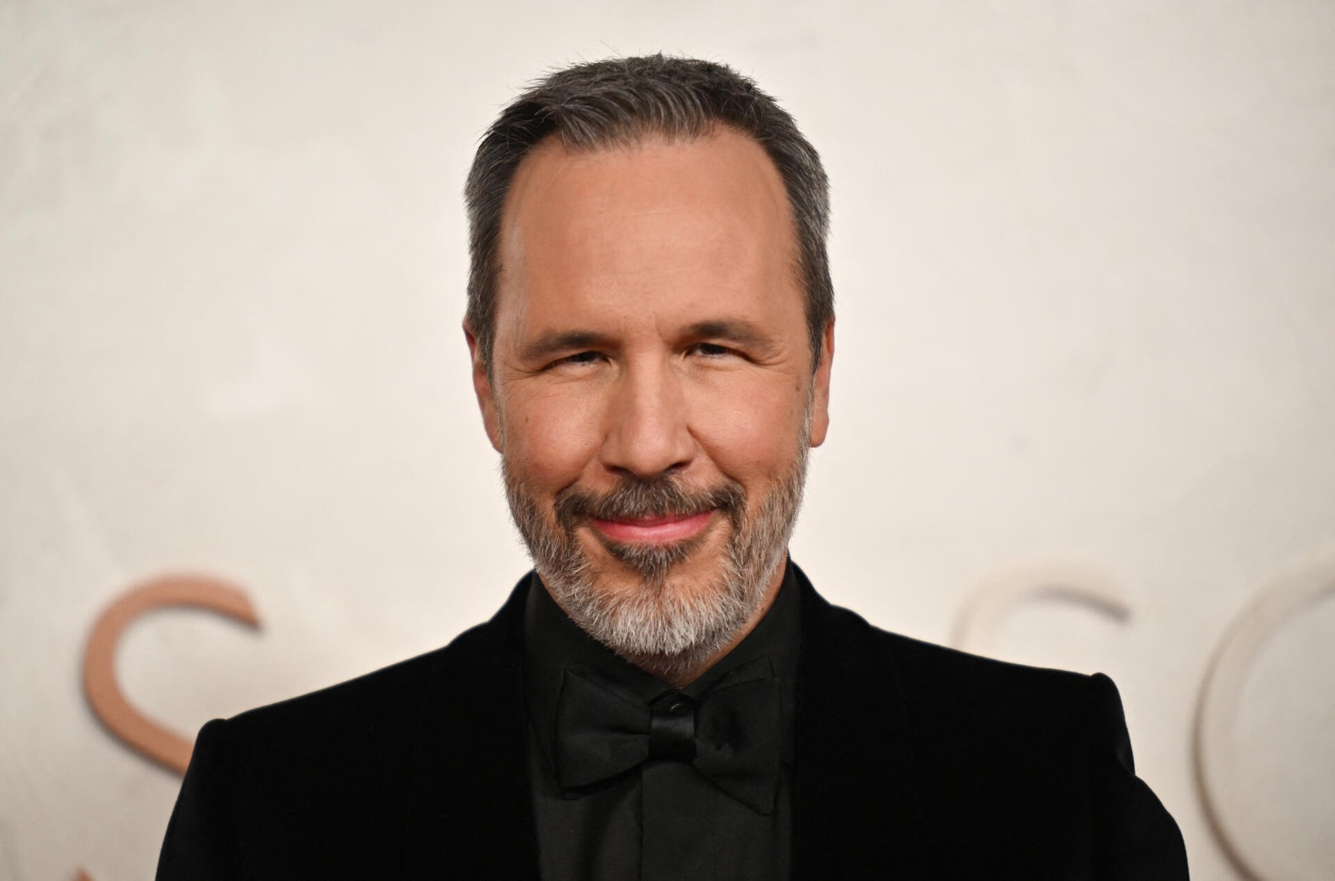 Denis Villeneuve smiles confidently at the Academy Awards, captured in a 4K Ultra HD image, showcasing his stylish appearance against a blurred background.