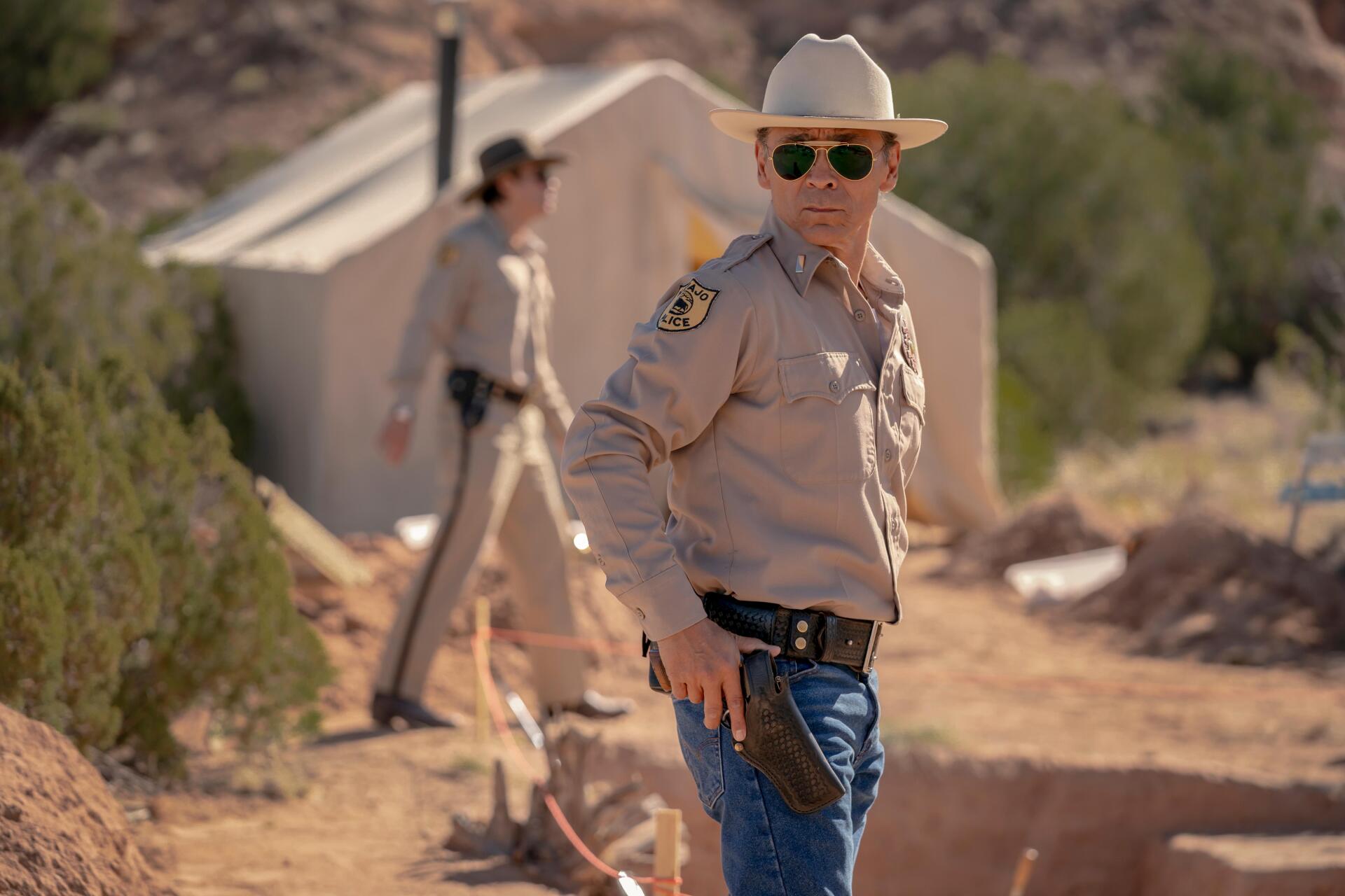 A scene from the TV show Dark Winds, featuring Zahn McClarnon in a sheriff's uniform, exuding a mix of authority and calm against a desert backdrop. 4K Ultra HD wallpaper quality.