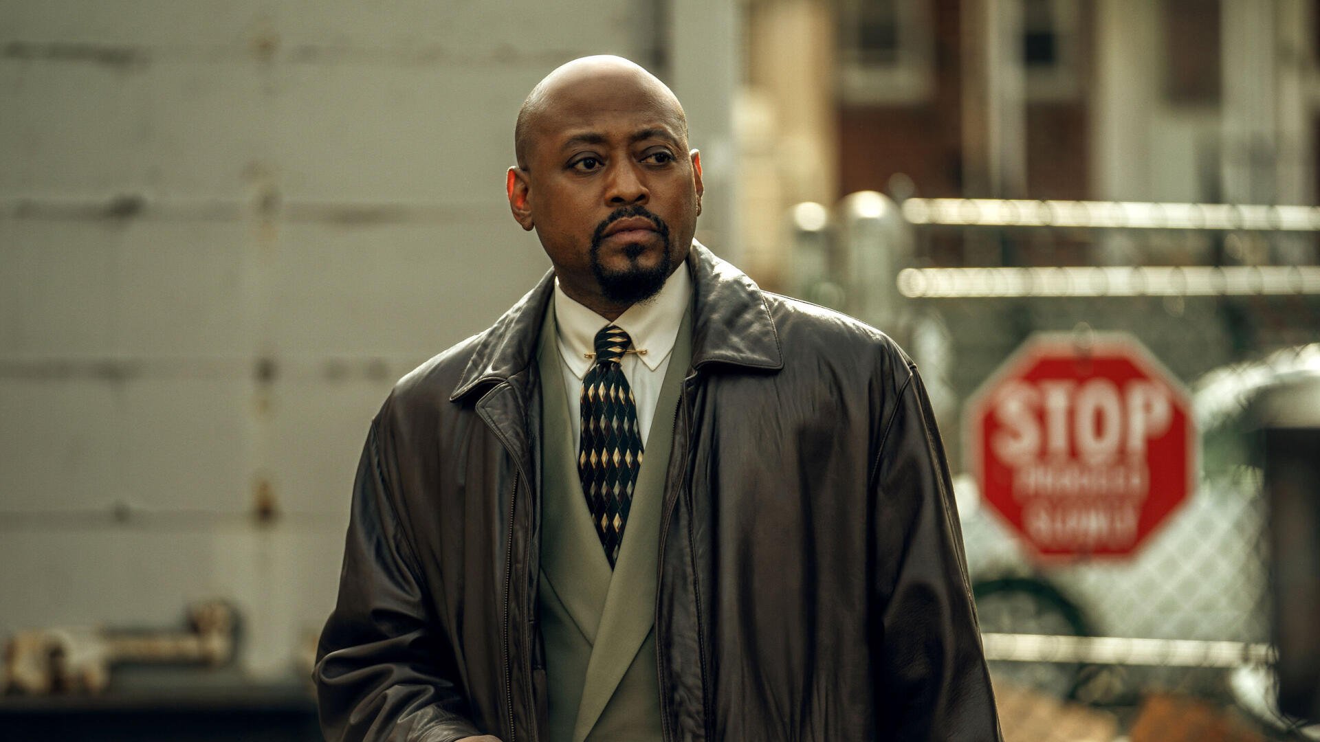 A character from the TV show Power Book III: Raising Kanan stands confidently, dressed in formal attire, set against an urban backdrop with a stop sign visible in the background.