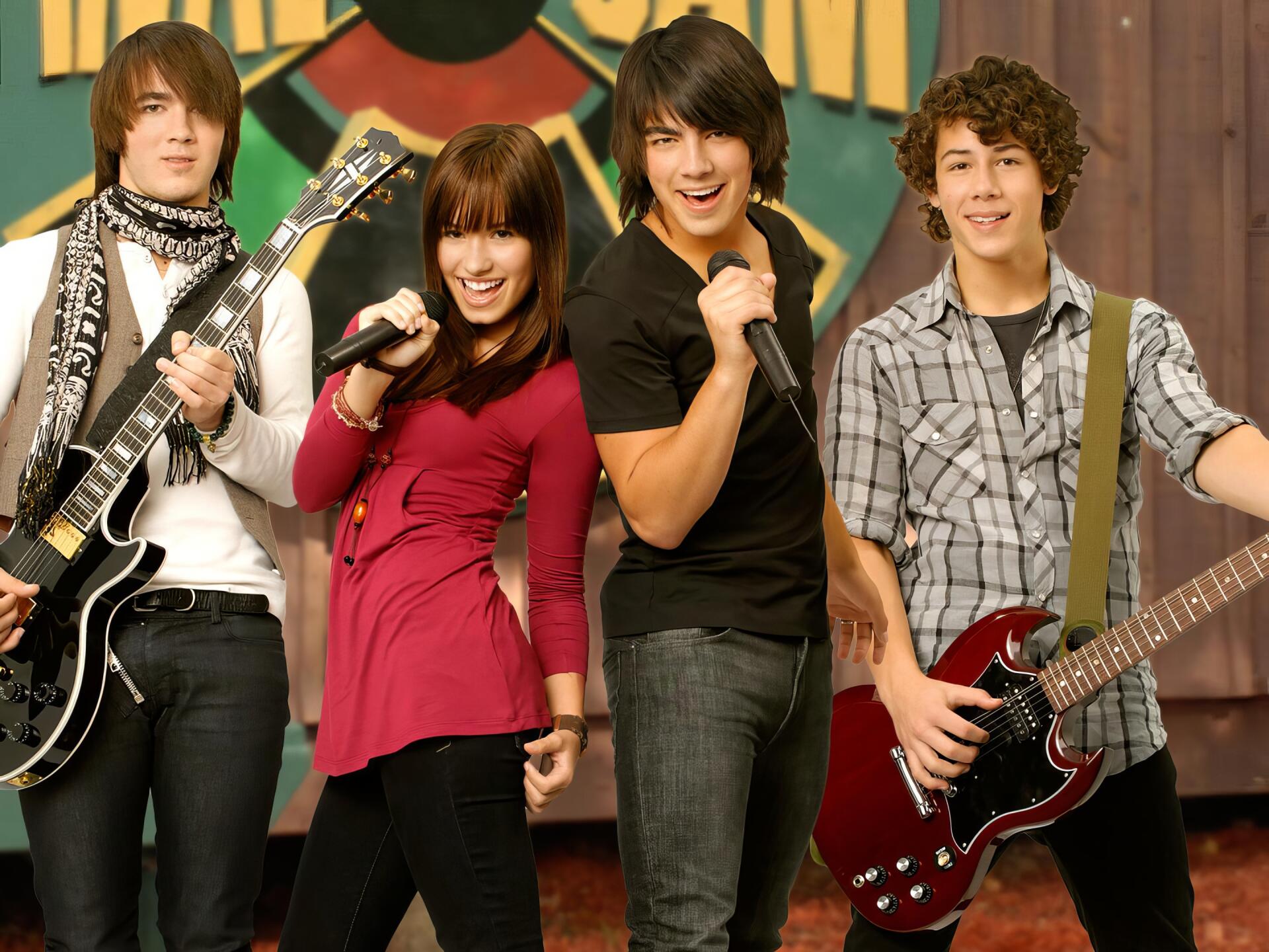 HD PC desktop wallpaper of the Camp Rock movie: four young musicians onstage—two guitarists, a singer with a mic and a backing vocalist—striking an upbeat band pose against a colorful backdrop.