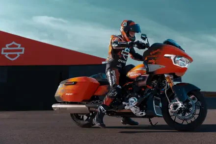 A rider in a racing suit poses on a vibrant orange Harley-Davidson CVO Road Glide RR motorcycle, set against a backdrop featuring the Harley-Davidson logo. 