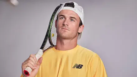 HD desktop wallpaper featuring tennis player Tommy Paul holding a racket, wearing a yellow New Balance shirt and white cap against a gray background.