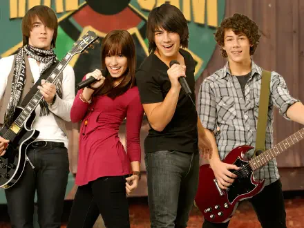 HD PC desktop wallpaper of the Camp Rock movie: four young musicians onstage—two guitarists, a singer with a mic and a backing vocalist—striking an upbeat band pose against a colorful backdrop.