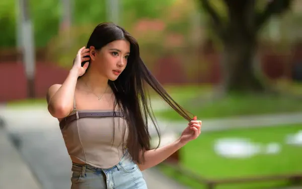 8K Ultra HD PC wallpaper: young woman in a tube top and jeans brushes her long hair, standing against a softly blurred green park background.