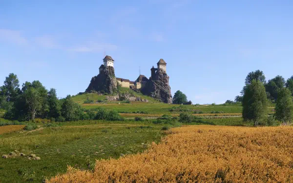 A stunning 4K Ultra HD landscape from Kingdom Come: Deliverance II, showcasing a medieval castle atop a lush green hill under a clear blue sky, surrounded by golden fields.
