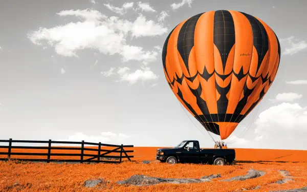 A vibrant orange hot air balloon ascends against a striking landscape, featuring a vintage truck and a wooden fence, creating an enchanting scene for a dream vacation backdrop.