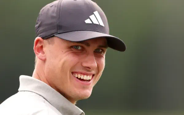 A close-up of Ludvig Åberg smiling while wearing an Adidas cap, set against a blurred green background. This HD image captures the energy and spirit of sports and golf.