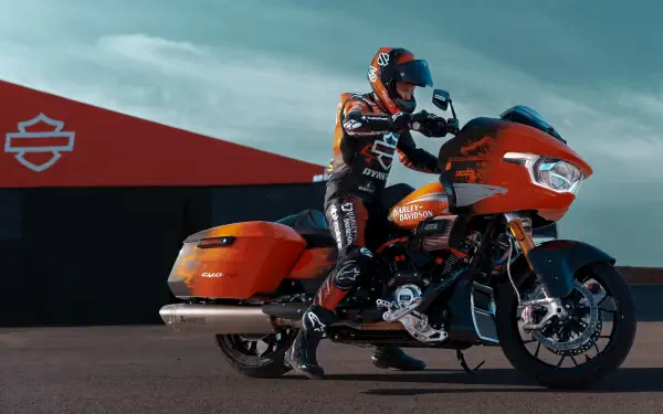 A rider in a racing suit poses on a vibrant orange Harley-Davidson CVO Road Glide RR motorcycle, set against a backdrop featuring the Harley-Davidson logo. 