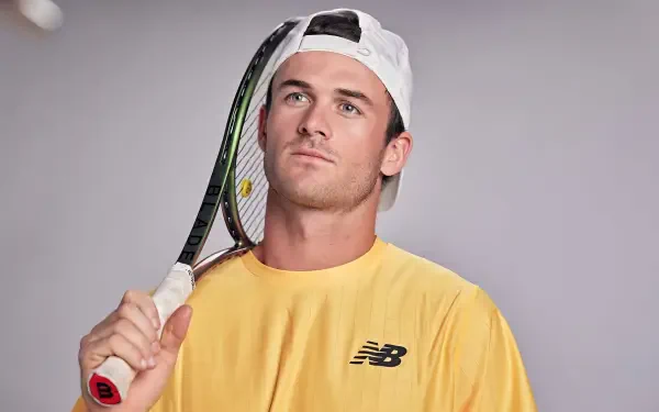 HD desktop wallpaper featuring tennis player Tommy Paul holding a racket, wearing a yellow New Balance shirt and white cap against a gray background.