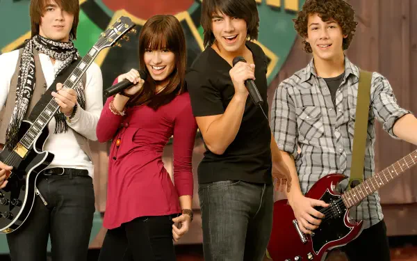 HD PC desktop wallpaper of the Camp Rock movie: four young musicians onstage—two guitarists, a singer with a mic and a backing vocalist—striking an upbeat band pose against a colorful backdrop.