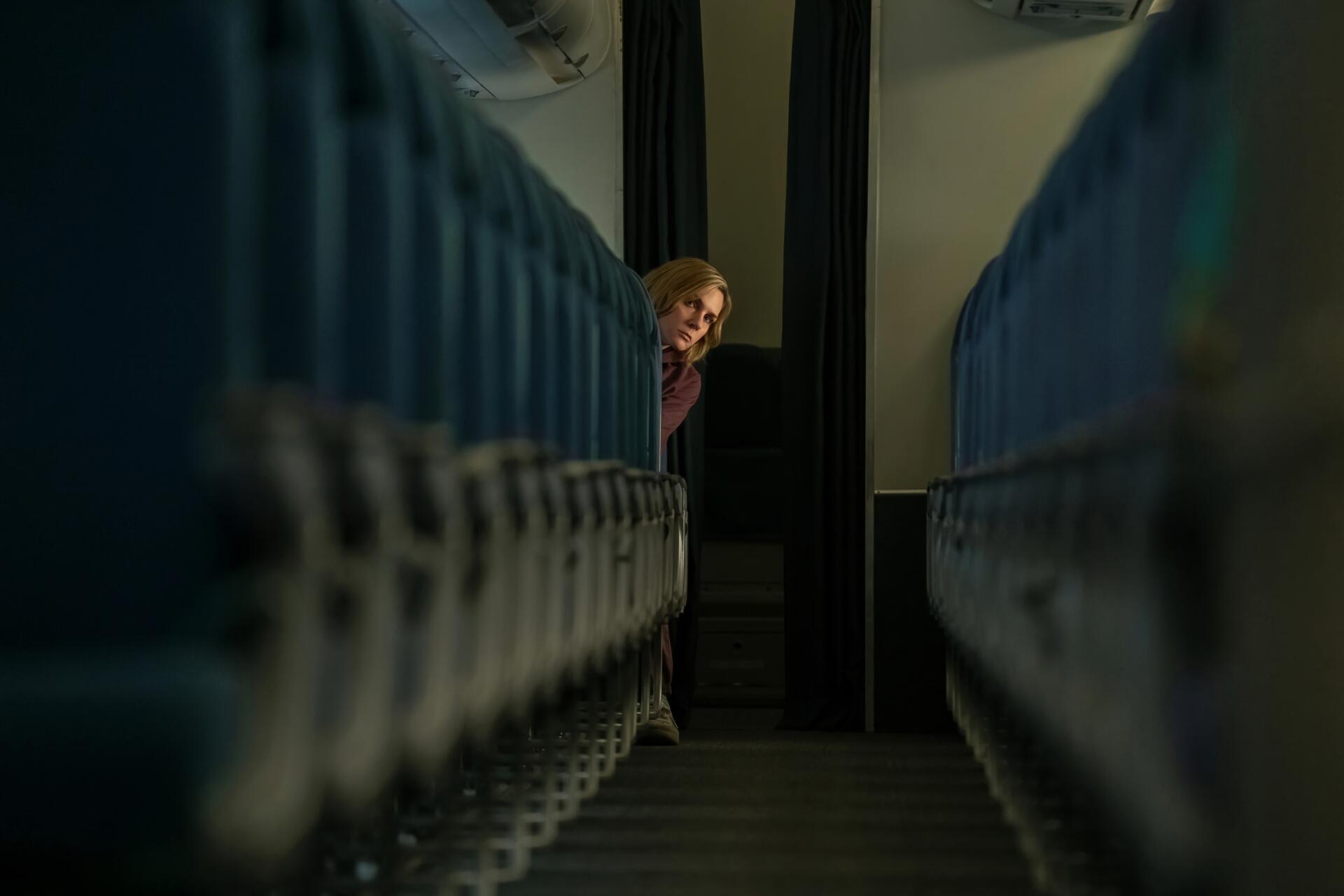 5K UHD desktop wallpaper from TV show Pluribus — a woman peeks between dim blue seats, framed by a narrow aisle.