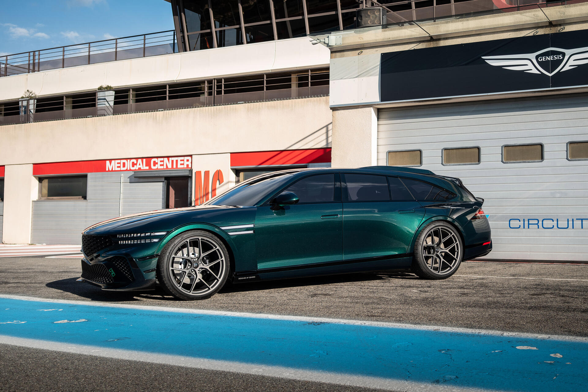 Side view of a teal Genesis G90 parked on a racetrack in bright sunlight — 2K Quad HD PC desktop wallpaper/background.