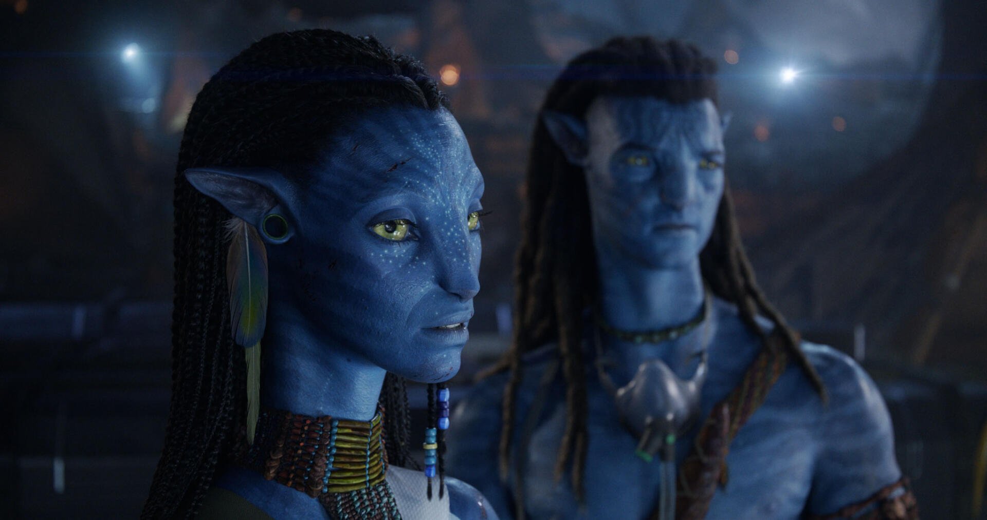 HD PC desktop wallpaper and background from the movie Avatar: Fire and Ash showing two blue-skinned Na'vi: a focused female with braided hair and jewelry, and a blurred male behind her.