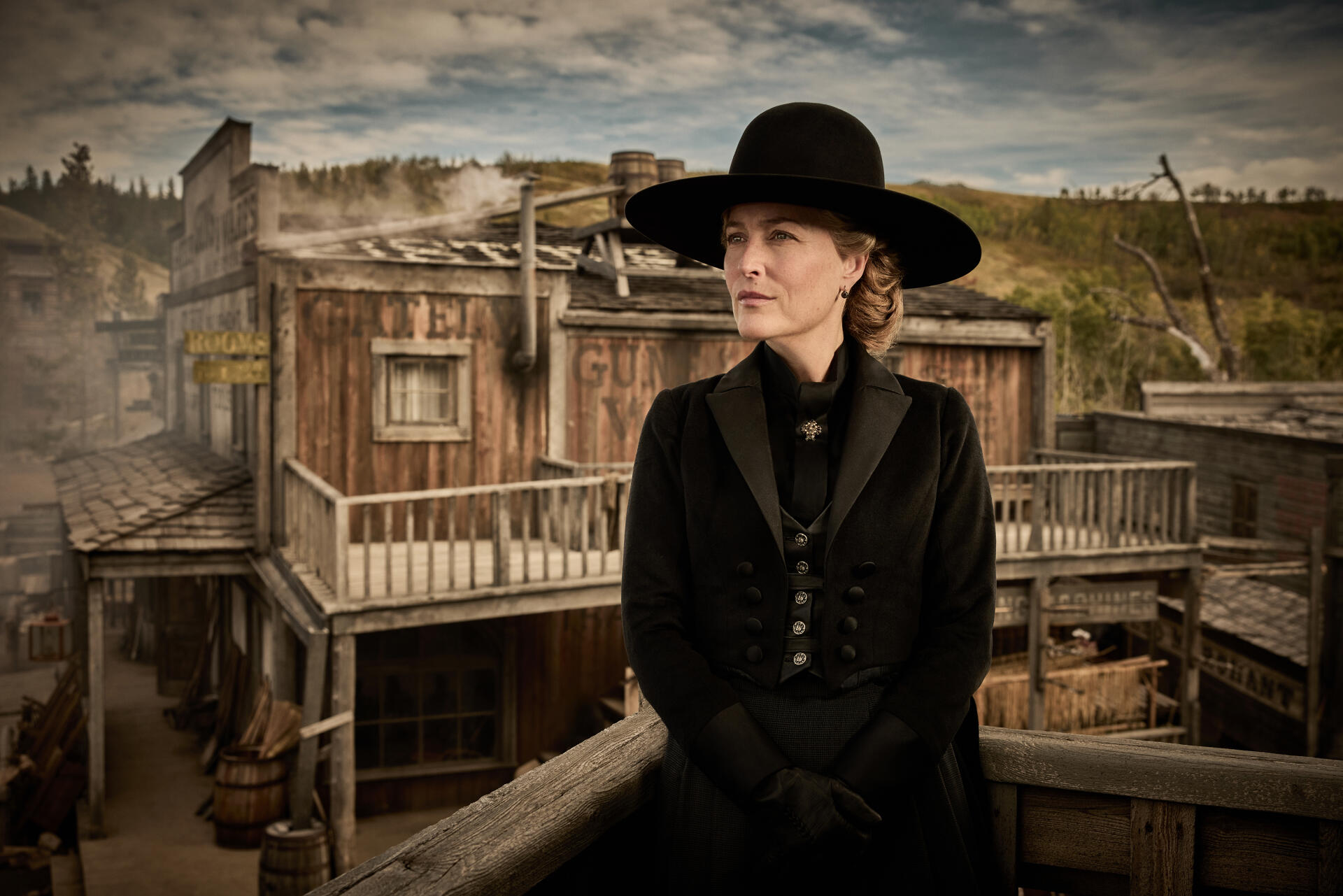 2K Quad HD PC wallpaper: a lone actress in a period black coat and wide-brim hat stands on a wooden balcony, gazing over a weathered Western town from the TV show The Abandons.