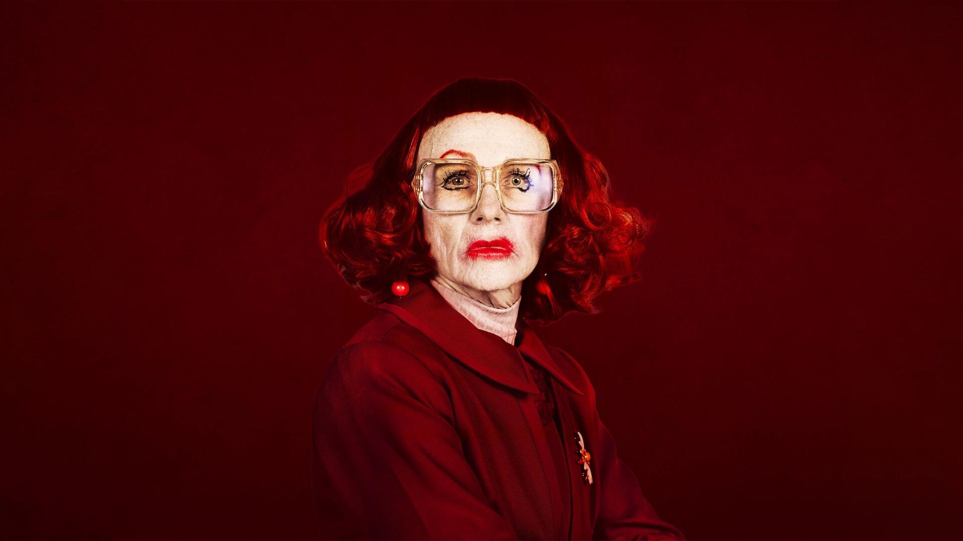 4K Ultra HD PC desktop wallpaper: portrait of an elderly woman with red hair and glasses against a deep red backdrop, weapon-themed, tense cinematic mood.