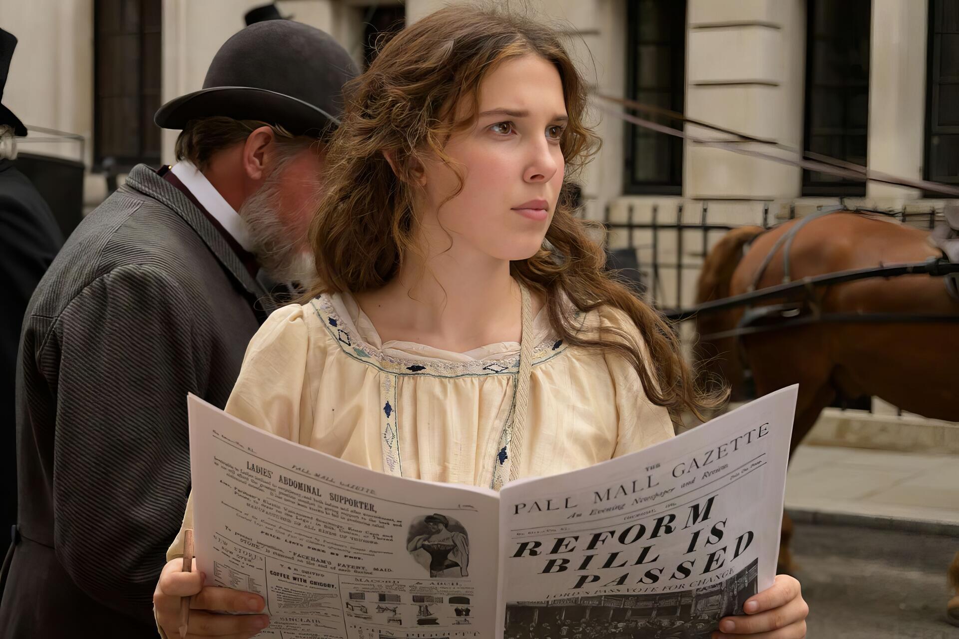 HD PC wallpaper from the movie Enola Holmes 3: young woman in Victorian dress reads a newspaper on a cobblestone street, horse-drawn carriage and period buildings in the background.
