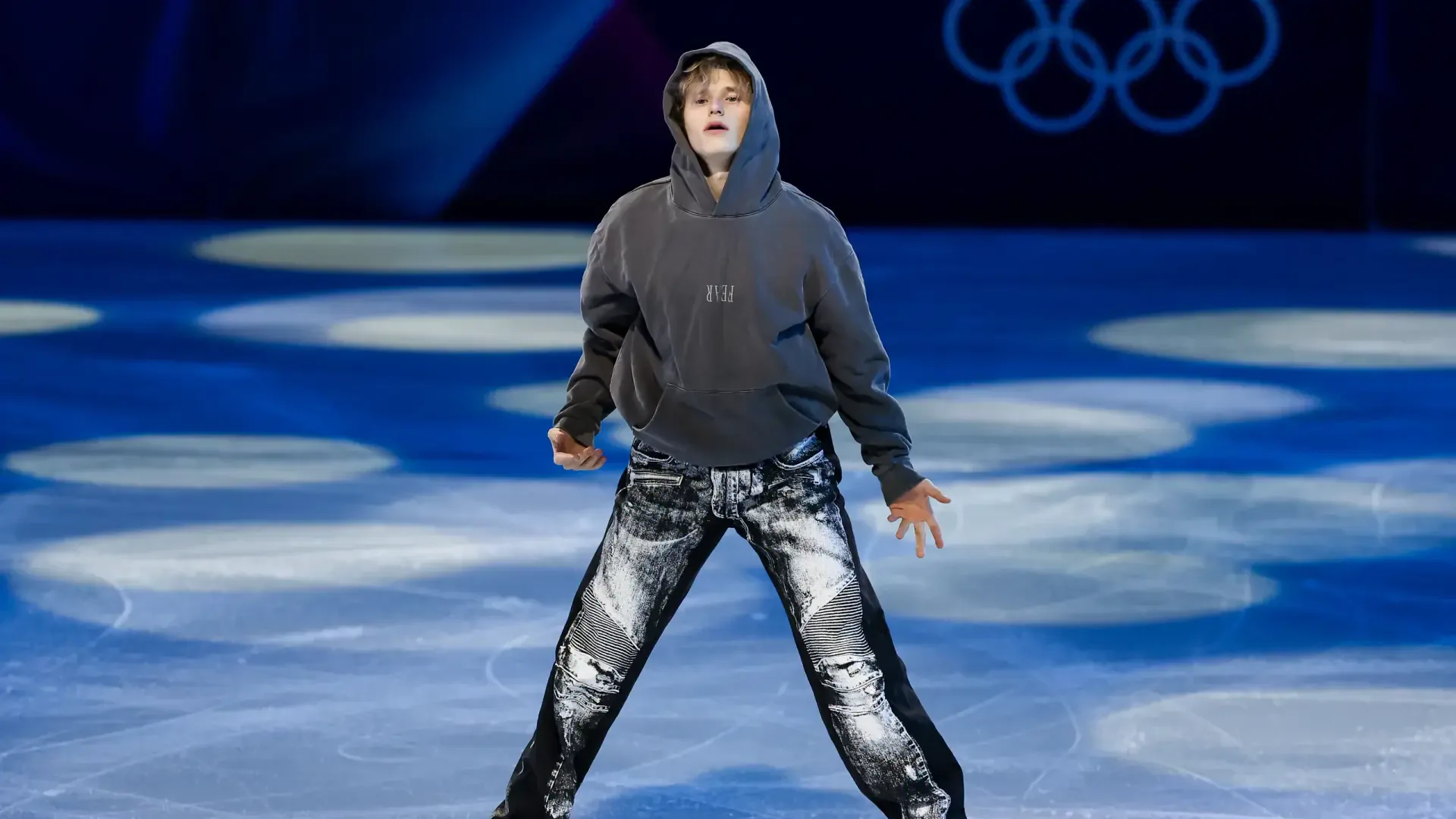 Figure skater in a hoodie and metallic pants performing on Winter Olympics ice beneath Olympic rings — 2K Quad HD PC desktop wallpaper