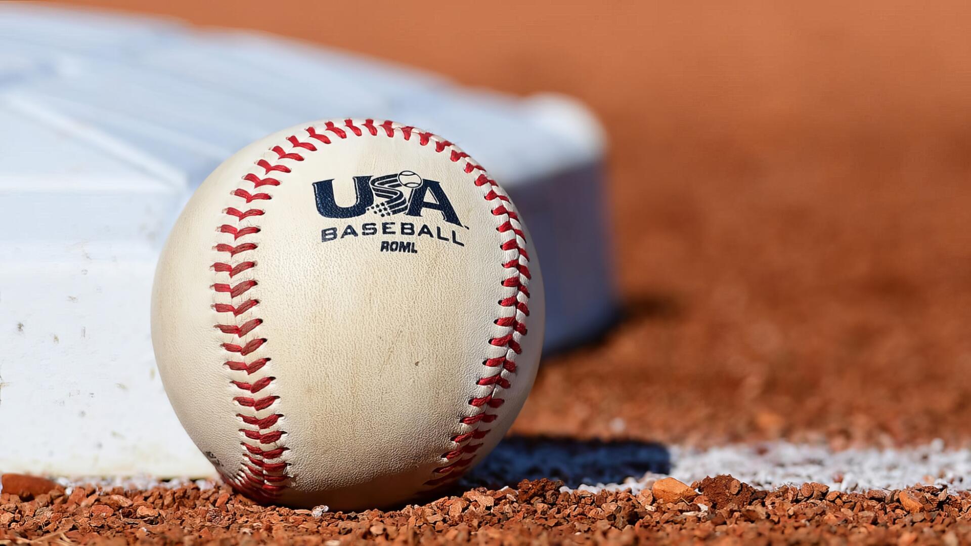 Close-up of a USA Baseball ball resting on infield dirt beside a base — sports-themed HD PC desktop wallpaper and background.