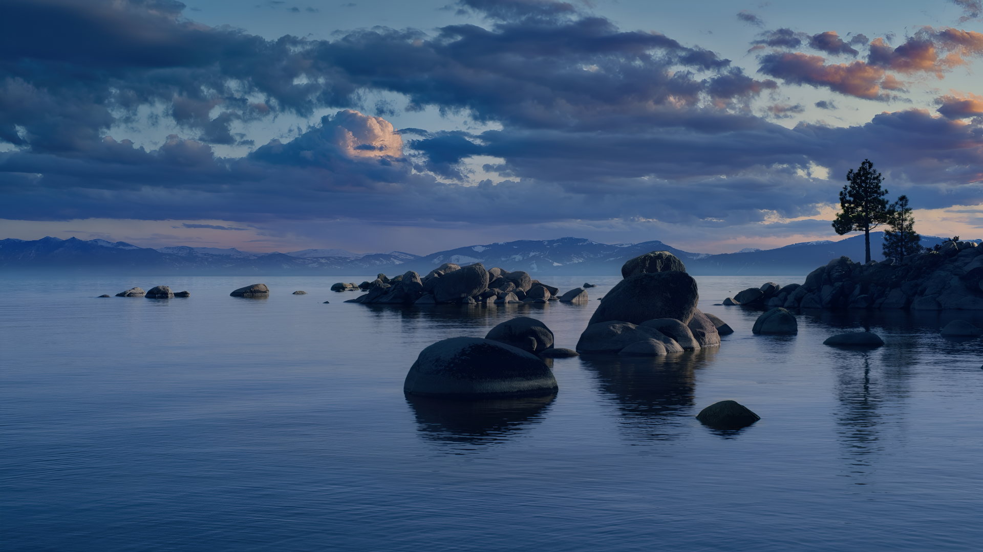 macOS 4K Ultra HD PC desktop wallpaper: calm lake landscape at dusk with glassy water, scattered boulders and distant trees beneath a soft, clouded sky.
