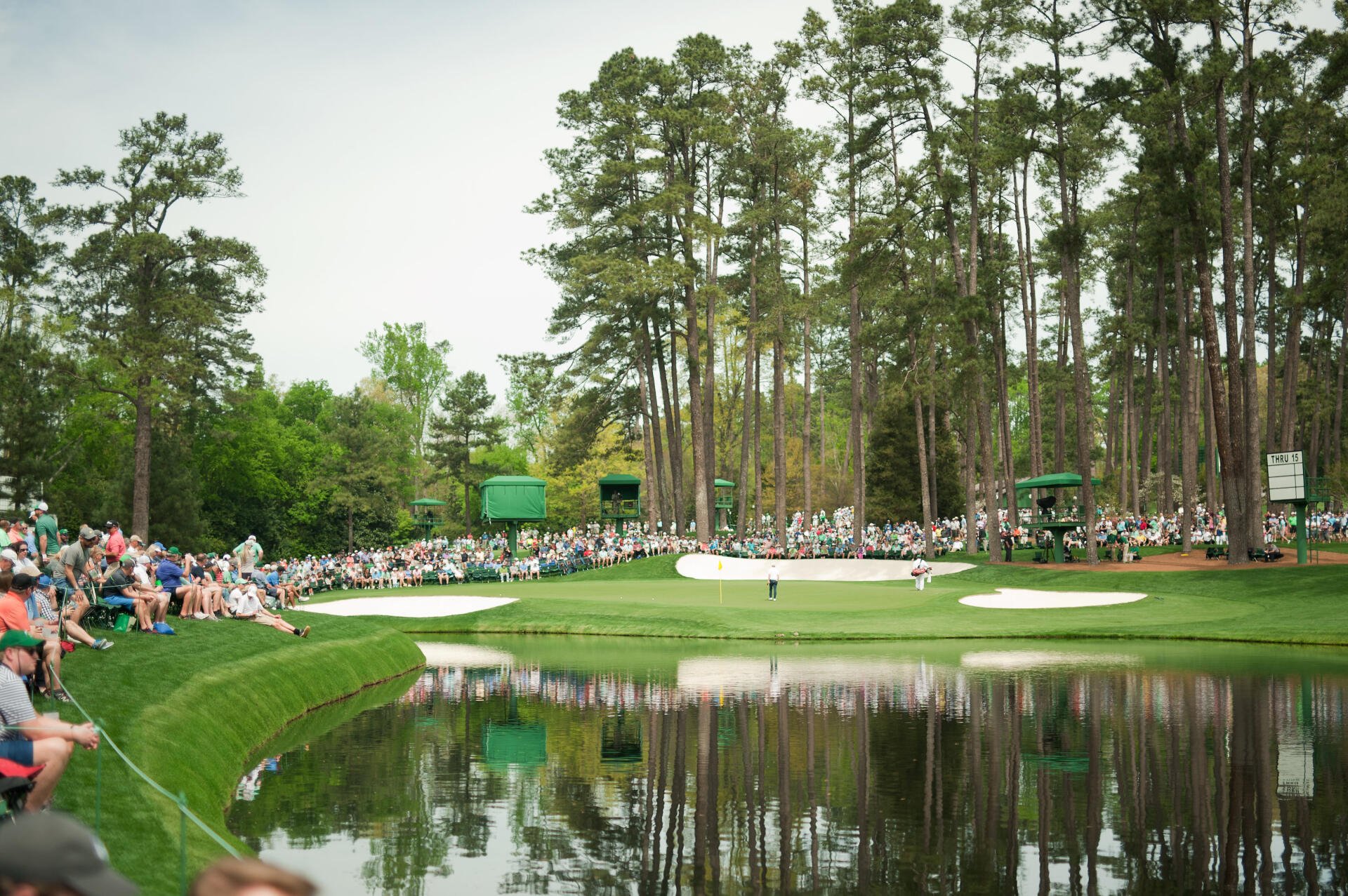 2K Quad HD PC desktop wallpaper — sports golf at The Masters: panoramic course view with green, water hazard, bunkers and spectators beneath tall pines.