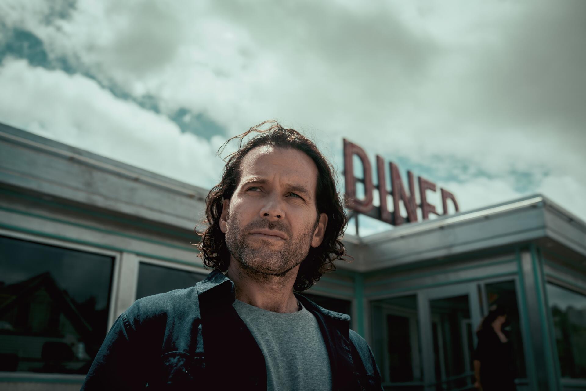 Long-haired man standing before a retro diner under stormy skies — TV show scene from 2022, presented as an 8K Ultra HD PC desktop wallpaper and background.
