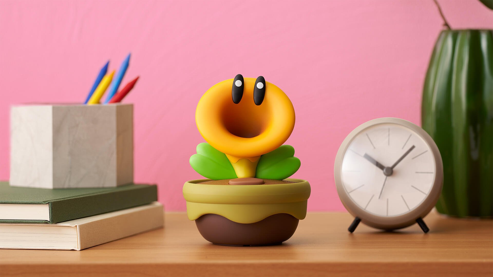 HD PC desktop wallpaper showing a Super Mario Bros. Wonder smiling yellow flower in a pot on a desk with books, pencils, an analog clock, and a pink wall background.