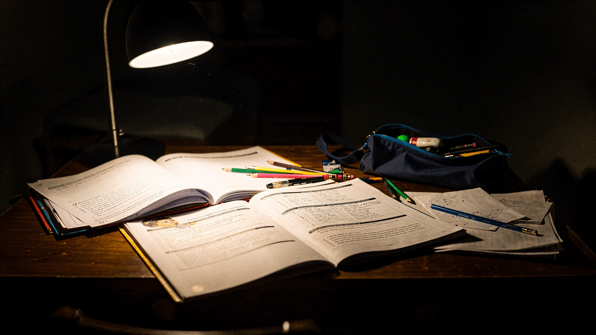 Homework scene: illuminated wooden desk with open notebooks, scattered pencils and a pencil case under a desk lamp — 4K Ultra HD PC desktop wallpaper and background.