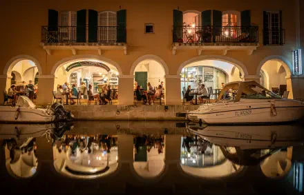 Côte d'Azur Mediterranean waterfront in France: arched restaurant terrace with diners and moored boats reflected in a calm canal — 5K Ultra HD PC desktop wallpaper.