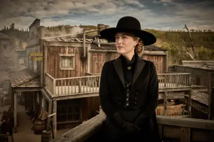2K Quad HD PC wallpaper: a lone actress in a period black coat and wide-brim hat stands on a wooden balcony, gazing over a weathered Western town from the TV show The Abandons.