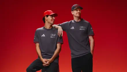 Two Audi F1 drivers in gray team shirts and red caps pose against a vivid red background — 4K Ultra HD PC desktop wallpaper.