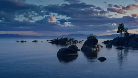 macOS 4K Ultra HD PC desktop wallpaper: calm lake landscape at dusk with glassy water, scattered boulders and distant trees beneath a soft, clouded sky.