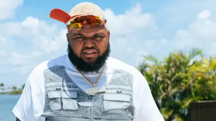 Close-up of an American comedian wearing a cap, sunglasses and a utility vest against a bright blue sky — 2K Quad HD PC desktop wallpaper/background.