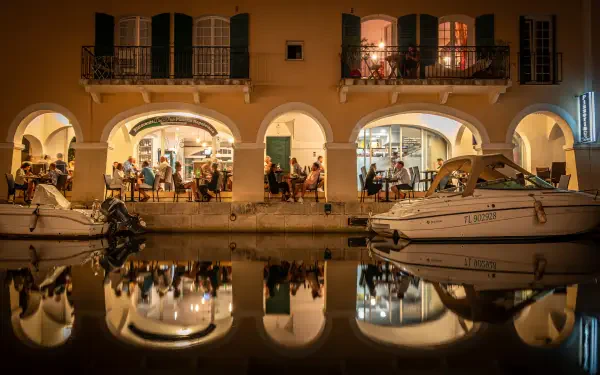 Côte d'Azur Mediterranean waterfront in France: arched restaurant terrace with diners and moored boats reflected in a calm canal — 5K Ultra HD PC desktop wallpaper.