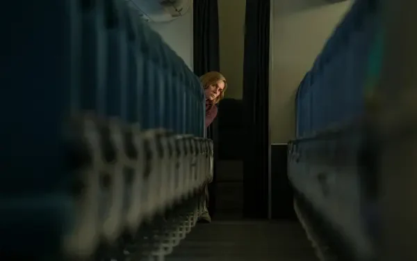 5K UHD desktop wallpaper from TV show Pluribus — a woman peeks between dim blue seats, framed by a narrow aisle.