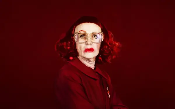 4K Ultra HD PC desktop wallpaper: portrait of an elderly woman with red hair and glasses against a deep red backdrop, weapon-themed, tense cinematic mood.