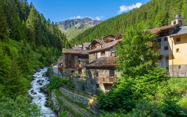 HD PC desktop wallpaper: a green mountain village beside a rushing forest stream, stone houses nestled in lush nature under tree-covered slopes.