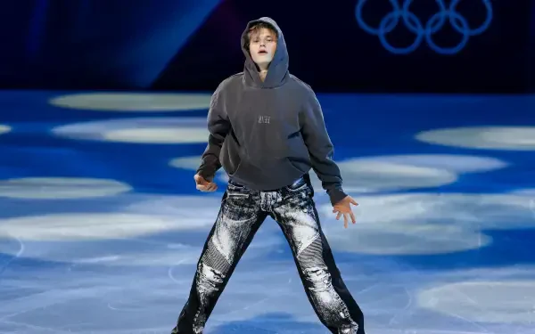 Figure skater in a hoodie and metallic pants performing on Winter Olympics ice beneath Olympic rings — 2K Quad HD PC desktop wallpaper