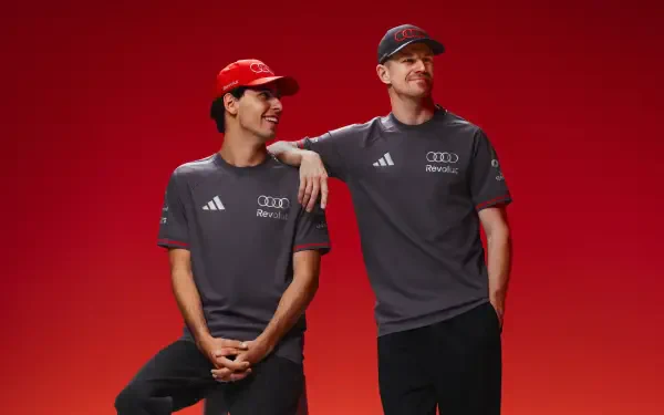 Two Audi F1 drivers in gray team shirts and red caps pose against a vivid red background — 4K Ultra HD PC desktop wallpaper.