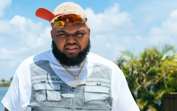 Close-up of an American comedian wearing a cap, sunglasses and a utility vest against a bright blue sky — 2K Quad HD PC desktop wallpaper/background.