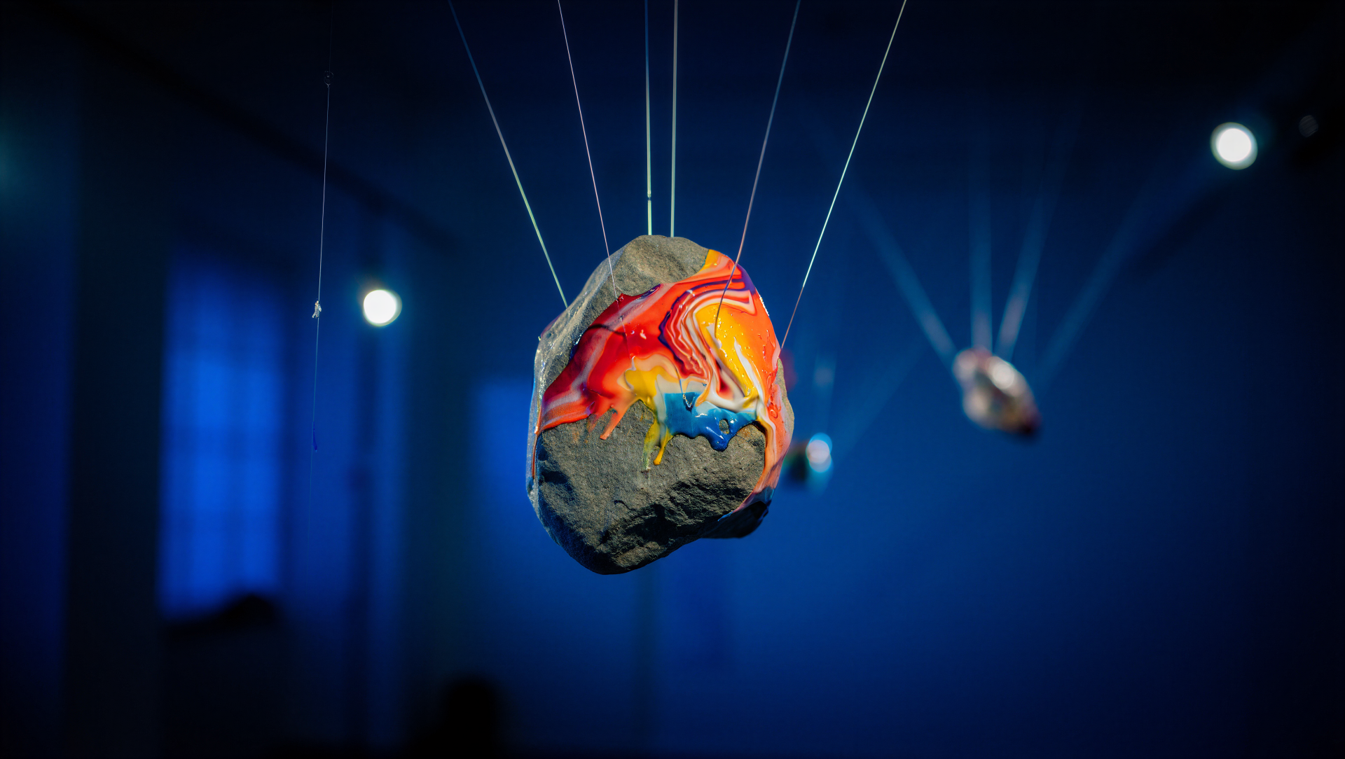 Rock painting: vivid red, yellow and blue swirls on a stone suspended by fine wires against a deep blue bokeh — 5K Ultra HD PC desktop wallpaper and background.