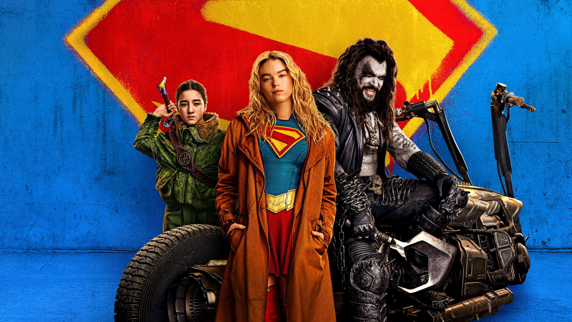 Supergirl (2026) 4K Ultra HD PC desktop wallpaper: Supergirl in blue-and-red costume with trench coat stands centered, flanked by allies and a motorcycle against a blue wall with S emblem.