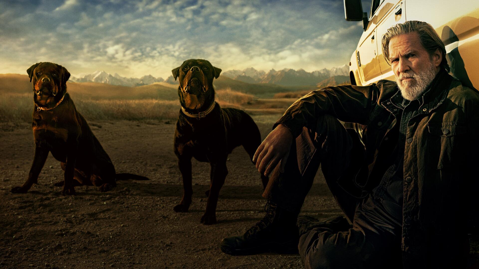 4K Ultra HD PC desktop wallpaper titled The Old Man of a rugged man seated by a vehicle with two dogs in a desert at dusk, moody skies and cinematic, rugged lighting.