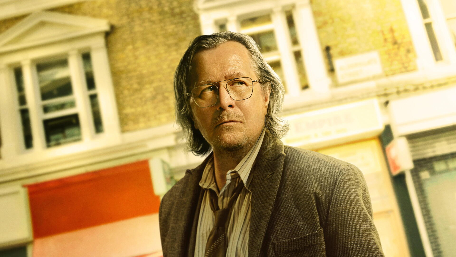 Slow Horses 4K Ultra HD PC desktop wallpaper and background: a pensive man with glasses and shoulder-length hair in a worn jacket stands before a sunlit brick storefront.