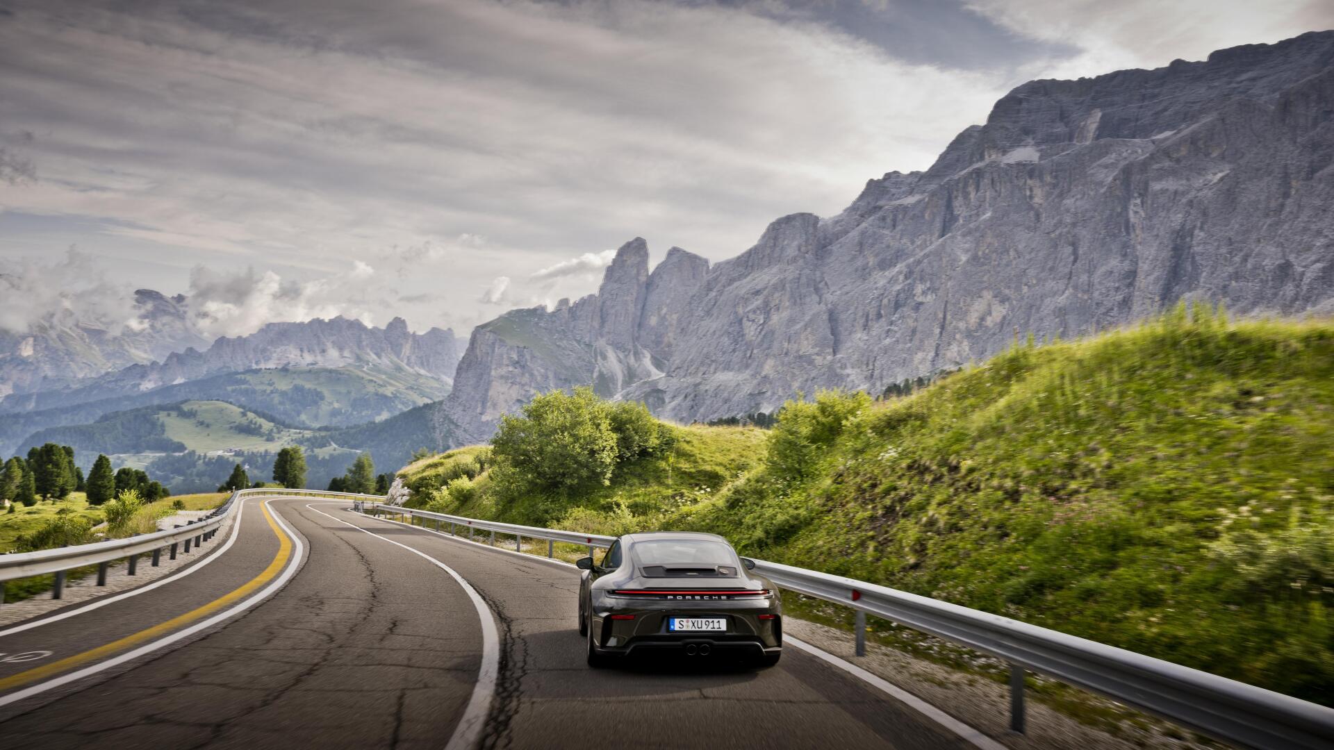 Porsche 911 GT3 on a winding mountain road with lush green hills and jagged peaks under a dramatic sky — 5K Ultra HD PC desktop wallpaper/background.