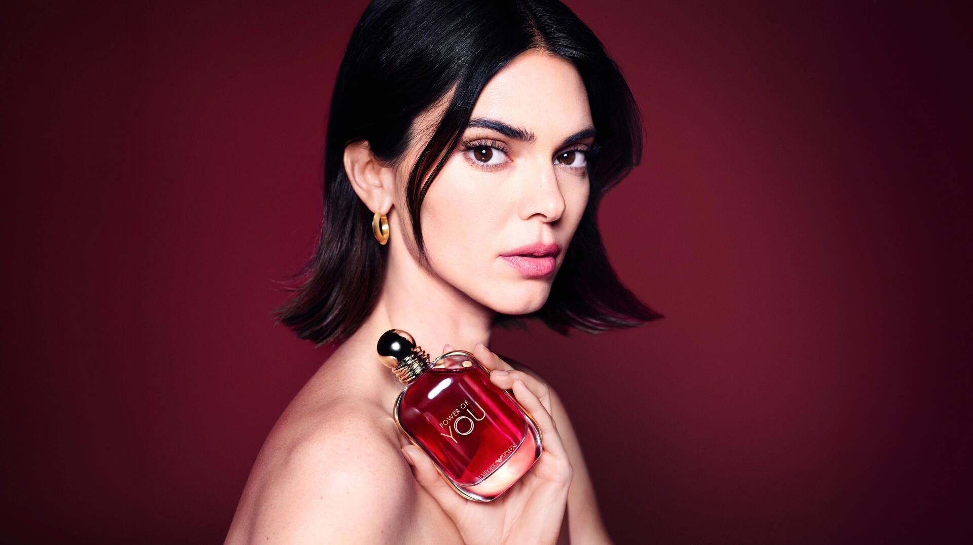 Dark-haired female model holding a red perfume bottle against a deep burgundy backdrop — 8K Ultra HD PC desktop wallpaper.
